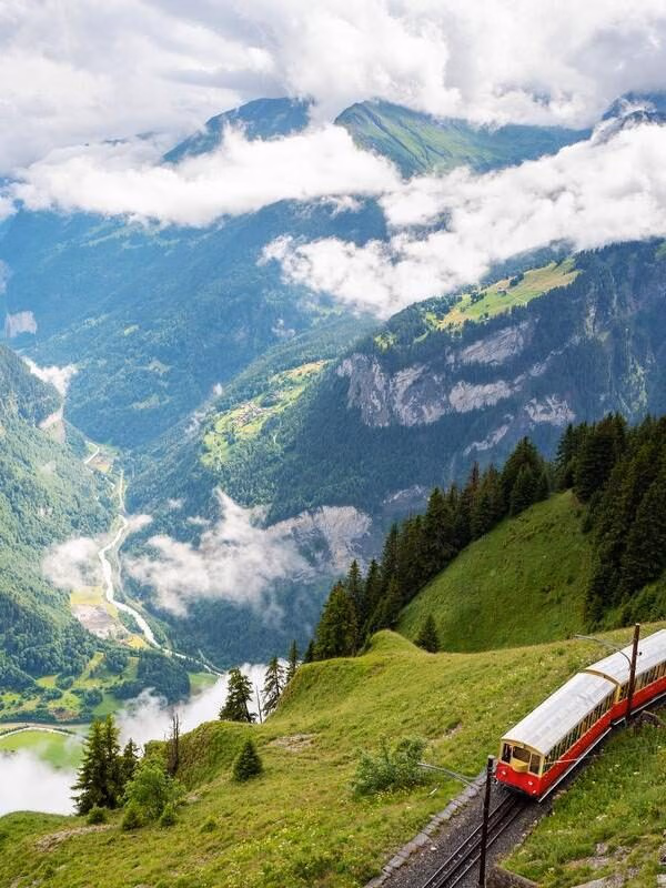 A red and white cogwheel train traveling up a lush green mountainside next to a deep valley filled with clouds.