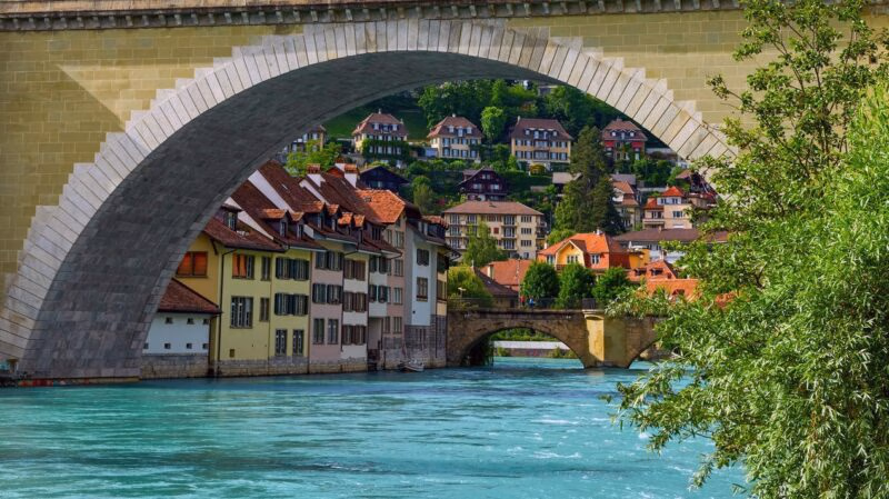 Historic waterfront buildings along a turquoise river seen through the stone arch of a large bridge in Bern.