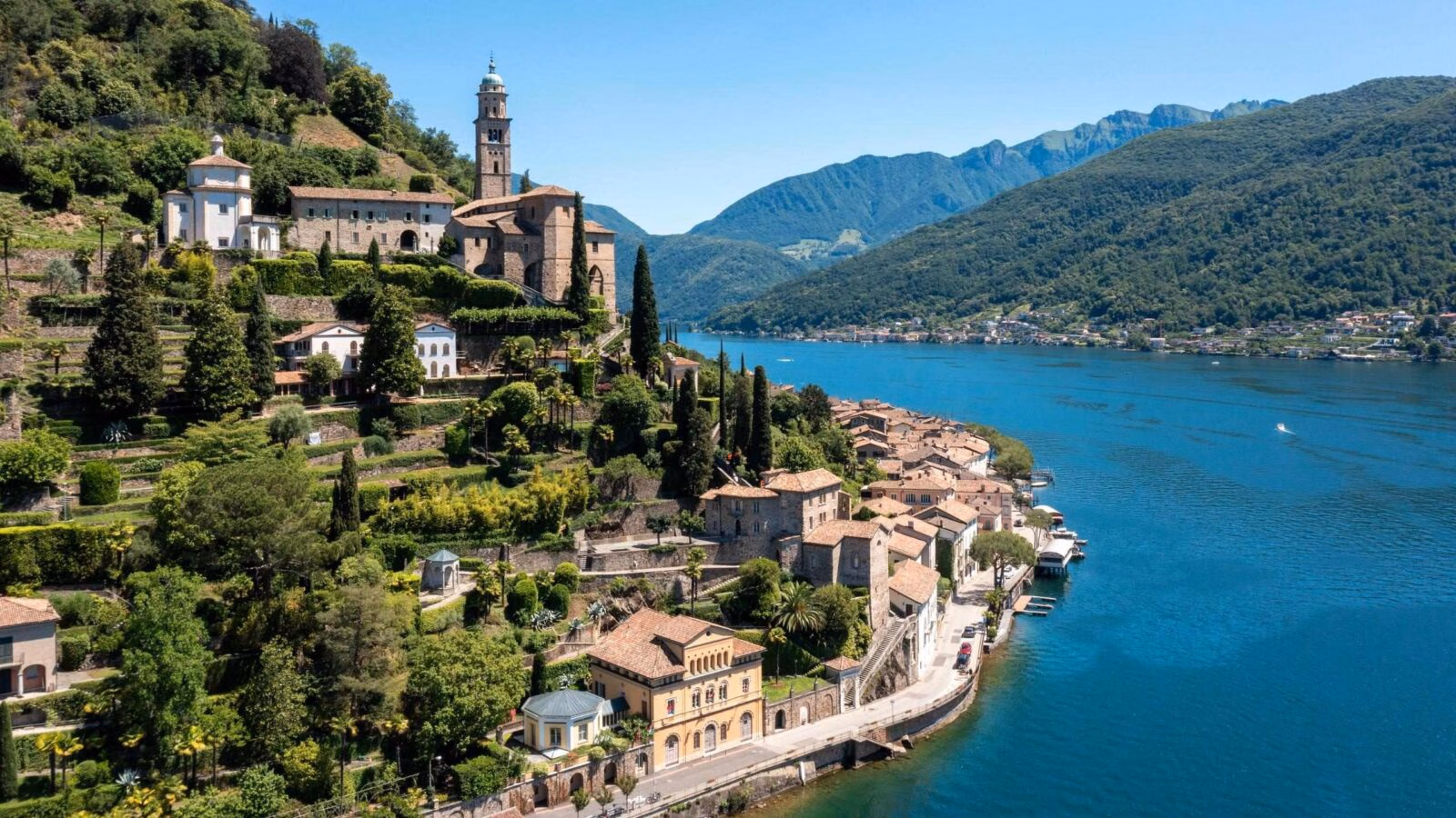A hillside village featuring historic stone buildings and a church spire alongside a large blue lake framed by mountains.