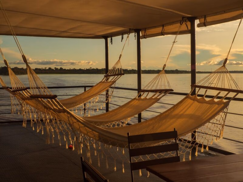 Three cream hammocks hang on a wooden boat deck overlooking a wide river at sunset on a latin america expedition cruise.