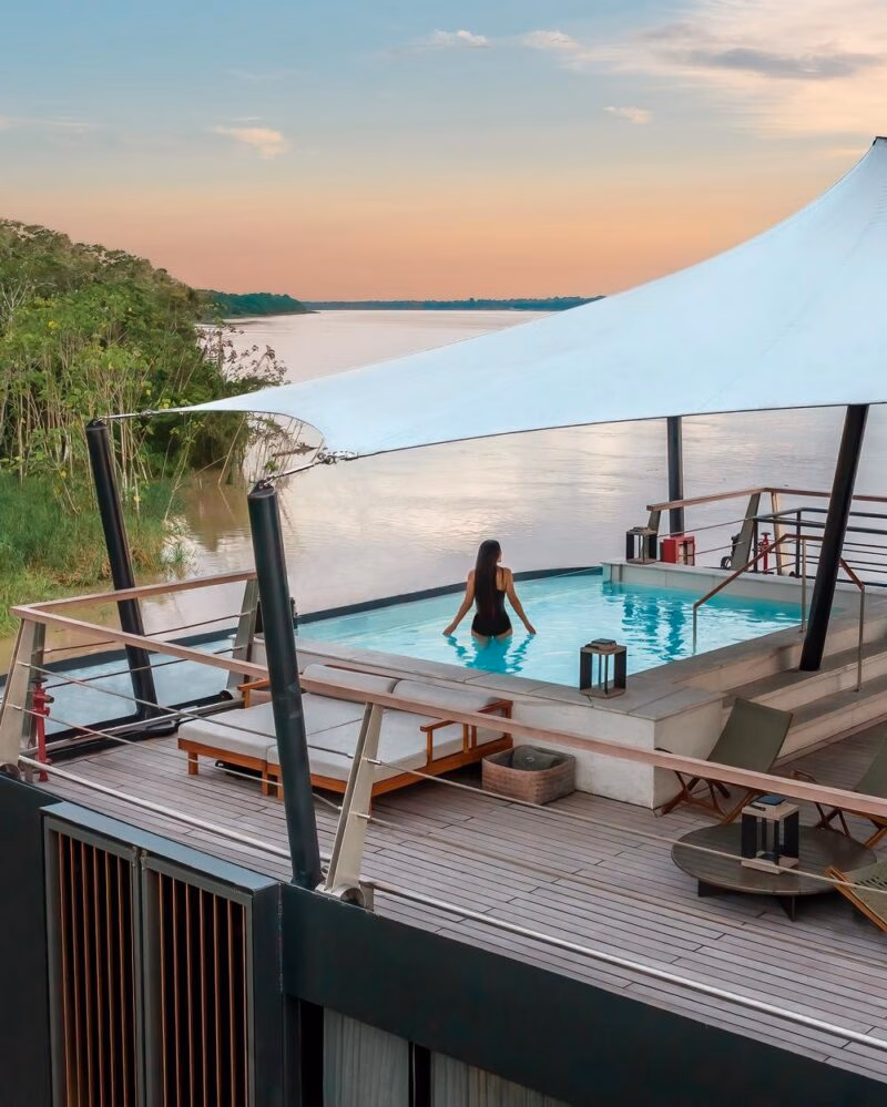 A woman stands in a shaded deck pool on a latin america expedition cruise, looking out at a wide river and green trees.