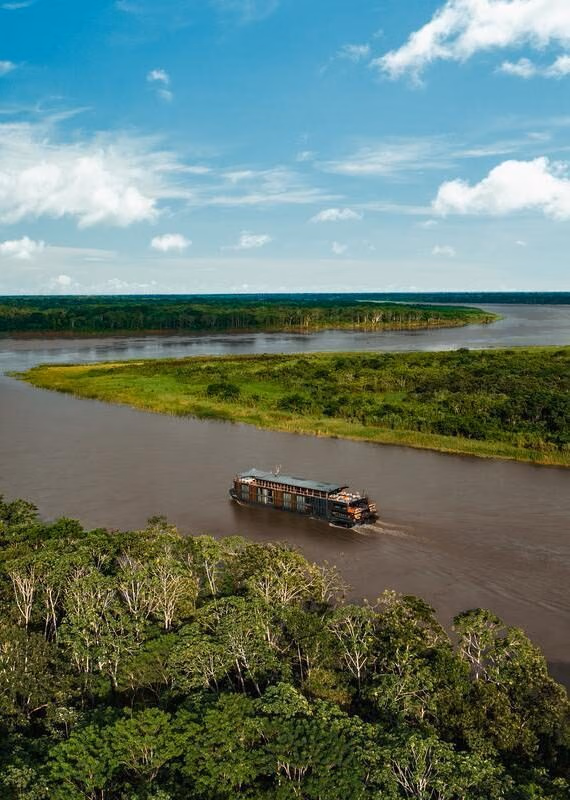 A modern latin america expedition cruise boat sails down a wide brown river surrounded by an expansive green rainforest.