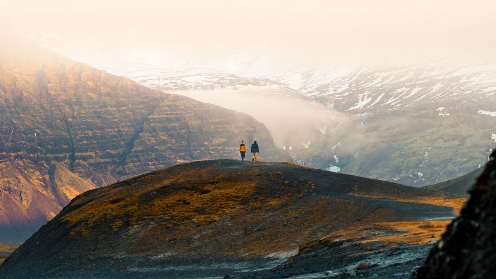 Distant view of two people walking on a dark hill ridge towards massive mountains and low-hanging clouds in a vast landscape.