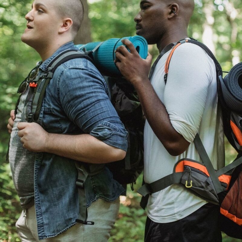 Two men in casual clothing stand in a forest, wearing large hiking backpacks with rolled sleeping mats attached.
