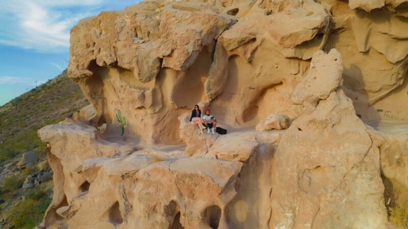Aerial view of a couple sitting in a cave-like niche of a large tan rock formation featuring a painted cactus and graffiti.