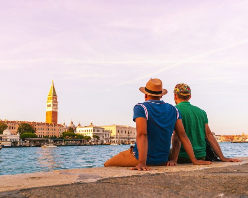Two men in hats sit on a stone ledge, looking across a wide canal at a historic city skyline with a tall bell tower.