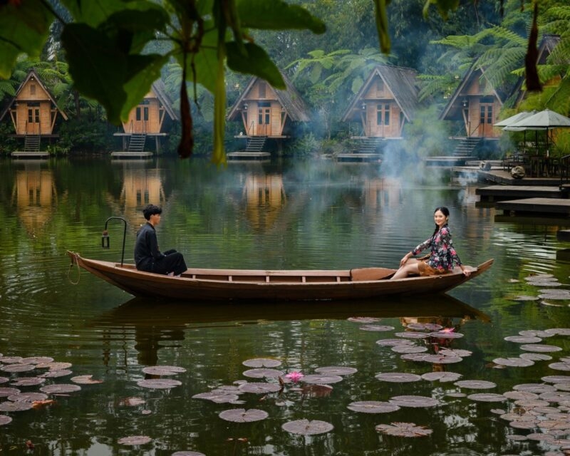 A man and a woman sit at opposite ends of a wooden boat floating on a calm lake lined with A-frame cabins on stilts.