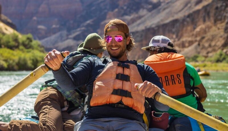 A smiling man in sunglasses and a life jacket rows a river raft. Another passenger's vest reads OARS.
