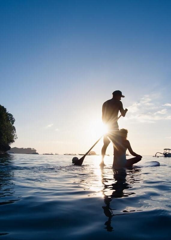 Silhouette of a standing man paddling and a sitting woman on a paddleboard on calm water against a bright sunburst.