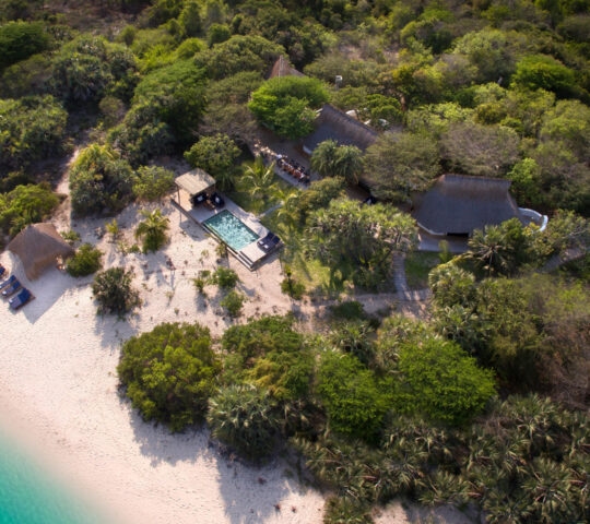 Aerial view of a private pool, thatched buildings, and white sandy beach at andBeyond Benguerra Island.