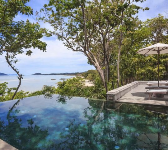 Infinity swimming pool with a scenic ocean view, surrounded by tropical trees and patio lounge chairs.