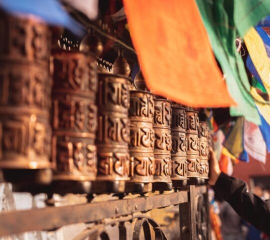 A row of bronze Buddhist prayer wheels beneath multi-coloured prayer flags in Nepal.