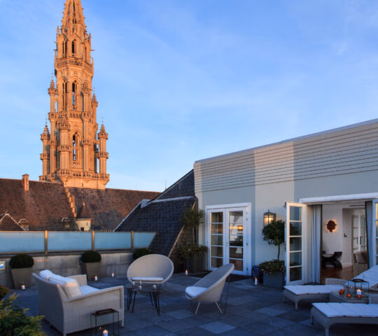 Rooftop terrace at Hotel Amigo Belgium with lounge seating, candles, and views of a tall gothic spire at dusk.