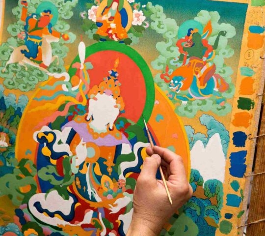 A close-up image of a half-finished Thangka painting and a hand holding a paintbrush.