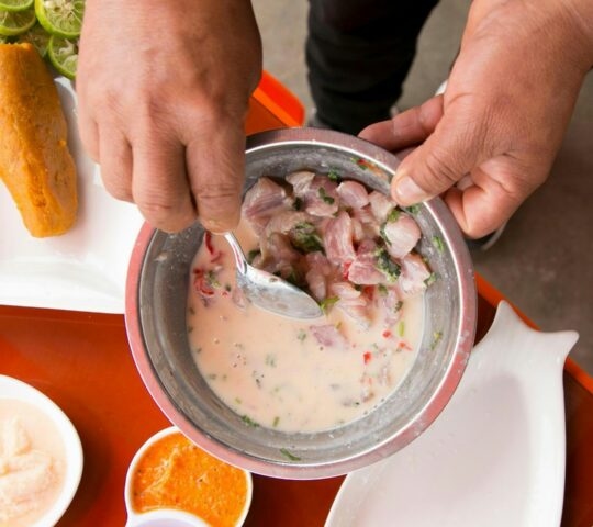 Hands mixing fish ceviche in a metal bowl on a lima tour, with sweet potato, limes, and toasted corn on an orange table.