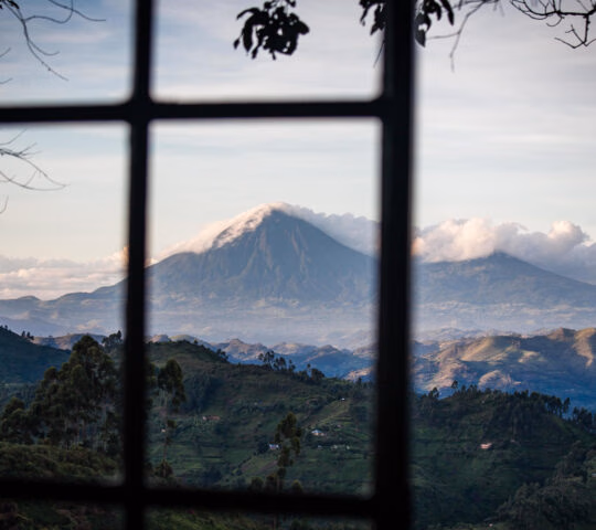 View through a window of a towering volcano and lush green hills at Clouds Lodge Uganda.