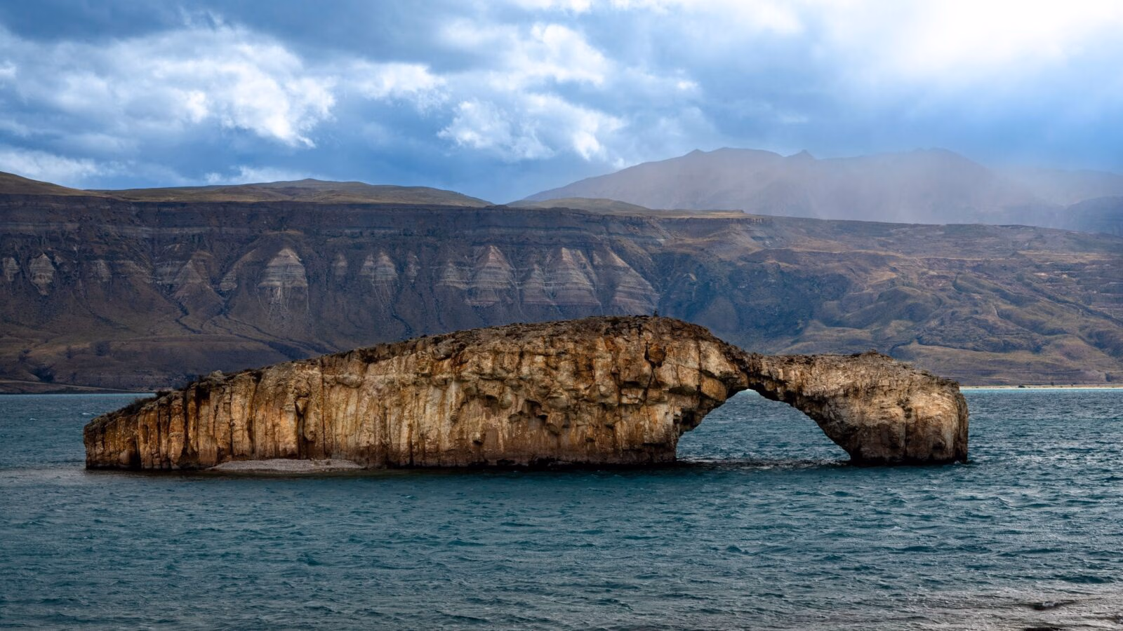 A large stone arch formation in a lake with mountains in the background in Posadas Argentina under a cloudy sky.