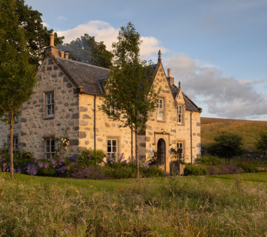 Stone farmhouse in mid distance with green meadow in the foreground and Scottish fells and blue sky beyond.