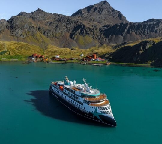 Large expedition cruise ship anchored in turquoise water near a shoreline with rusted buildings and rugged green mountains.