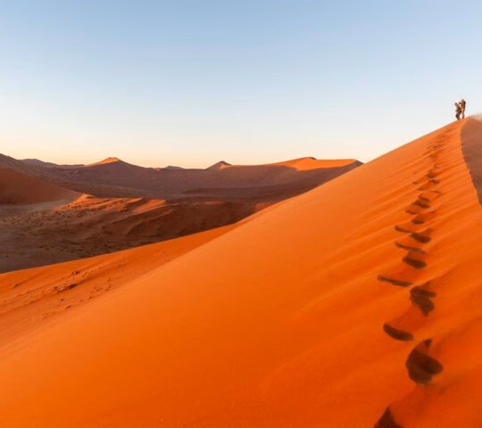 Footsteps leading up a huge red sand dune, with two people standing on top looking out over Sossusvlei desert