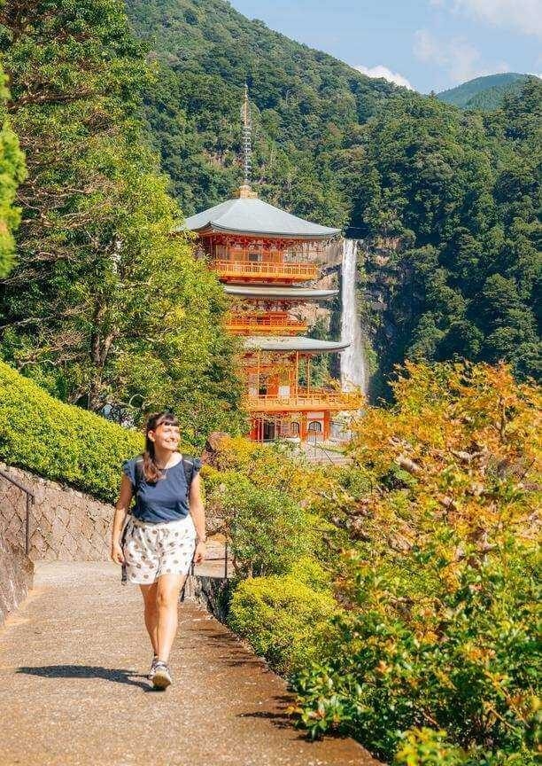 A smiling woman walks on a paved path with a three-story red pagoda and a tall waterfall in the background.