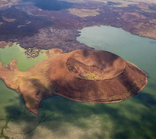 Aerial view of a large brown volcanic crater standing next to a green lake with rocky terrain.