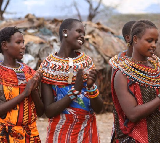 Four women wearing brightly coloured traditional attire and large beaded collars smiling together.