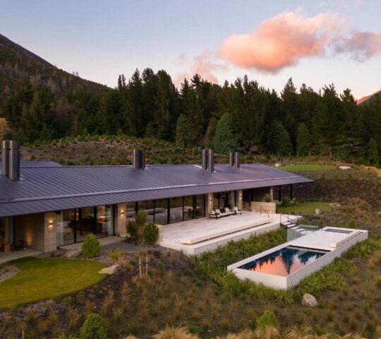 Aerial view of the Homestead at Flockhill Lodge, with a swimming pool, patio, and forest surrounded by mountains at sunset.