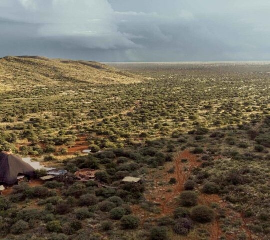 An aerial view of a thatched-roof lodge situated in a vast savanna landscape beneath cloudy skies.