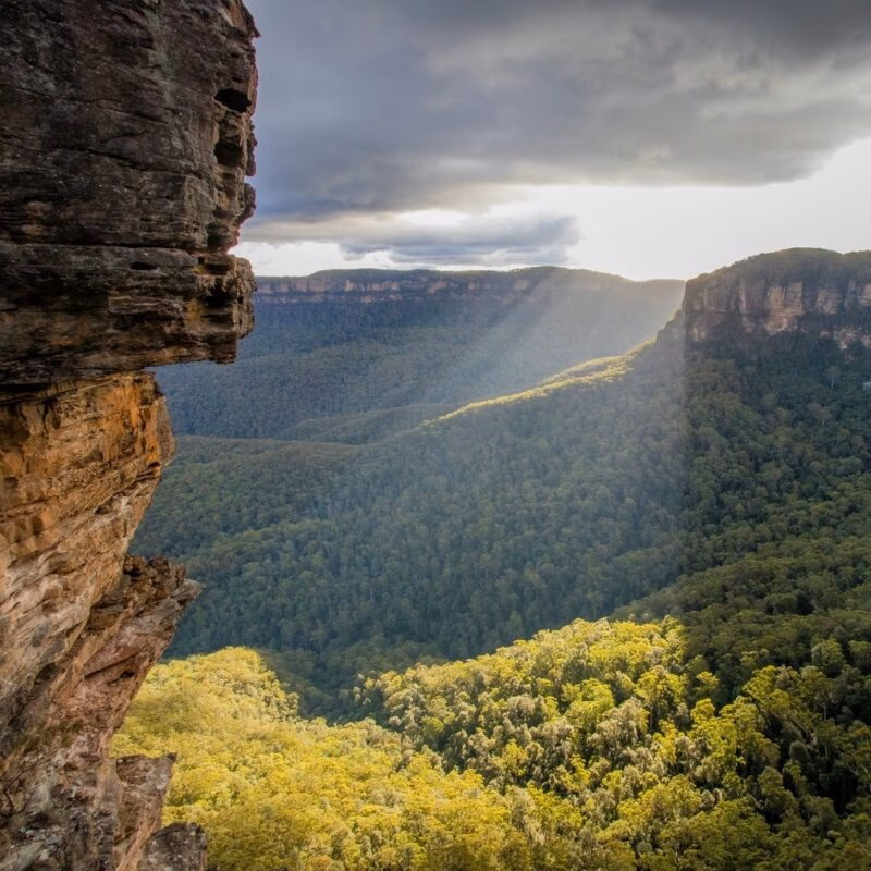 Sunlight beams through clouds over a vast, green forested valley and distant plateaus in Australia, framed by a rock face.