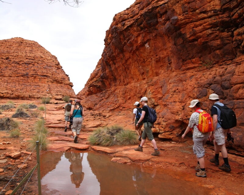 A group of hikers walking on a red dirt path between tall red rock walls in Australia.