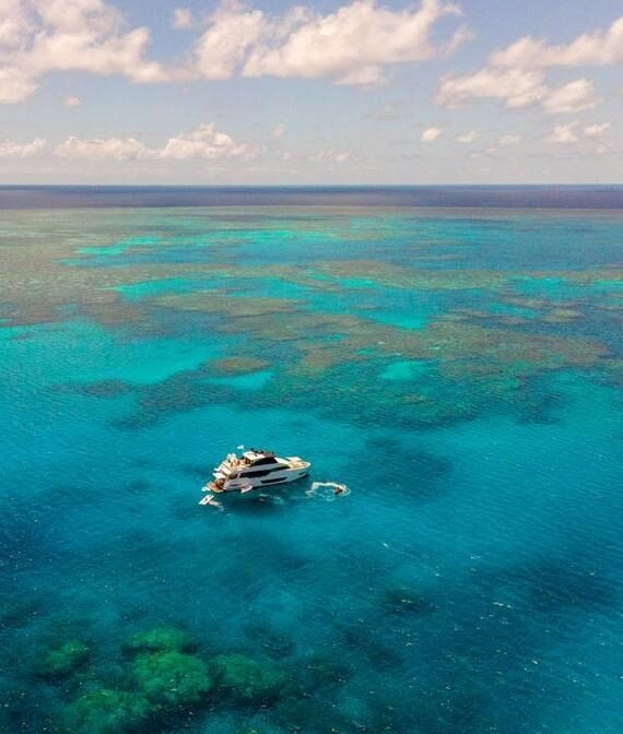 Aerial view of a white luxury yacht anchored in bright turquoise waters over a vast coral reef in Australia.