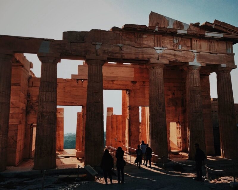 Silhouetted figures walking through the large stone columns of the ancient Acropolis gateway in Athens, Greece at sunset.