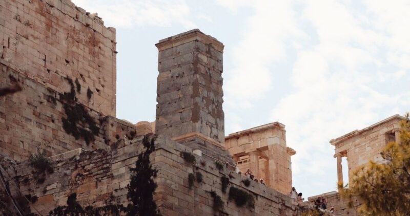 Visitors walking among the ancient stone ruins and tall pillars of the Acropolis in Athens, Greece under a cloudy sky.