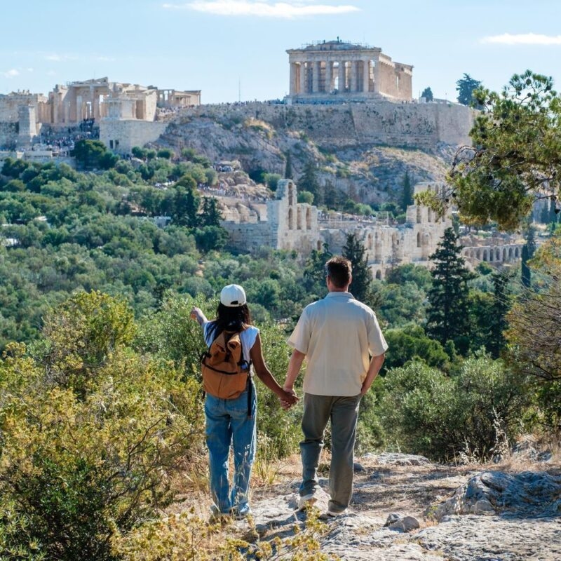 A couple holding hands on a nature trail, looking out over the ancient ruins of the Acropolis in Athens, Greece.