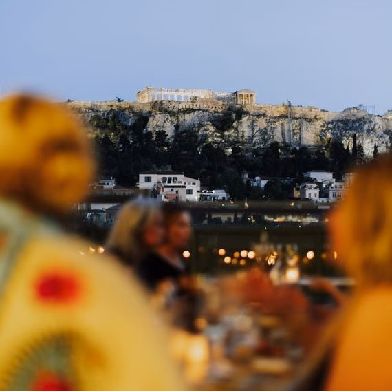Blurred figures at a dinner table in the foreground with a clear view of the lit-up Acropolis in Athens, Greece at dusk.