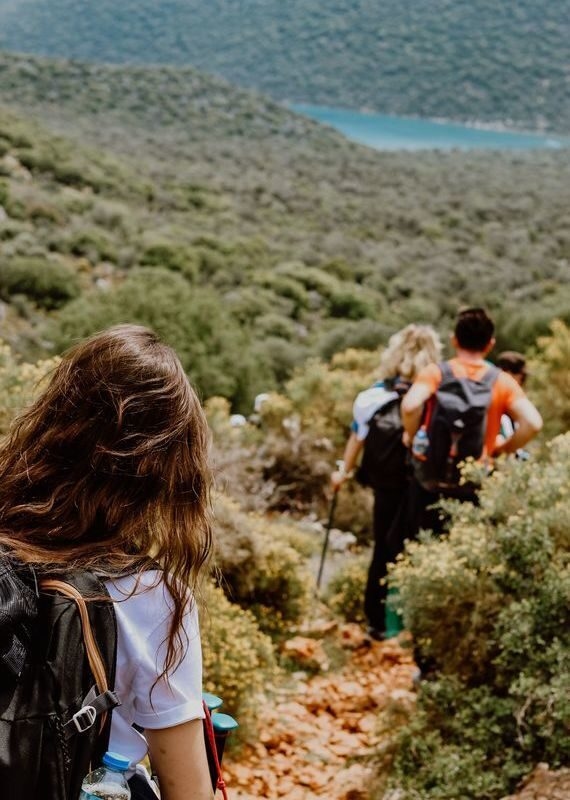 Group of people with backpacks hiking down a rocky dirt trail surrounded by green shrubs towards the sea in Greece.