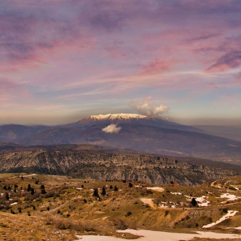 Landscape view of the snowy peak of Mount Olympus in Greece towering over rolling hills under a pink and purple sunset sky.