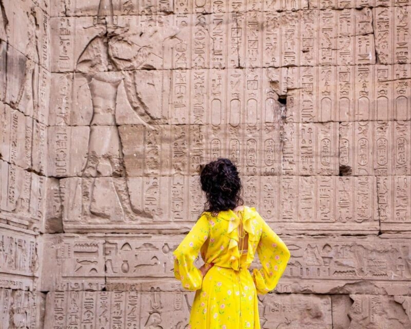 Woman in a yellow floral dress standing in front of a tall stone wall covered in ancient Egyptian hieroglyphs.