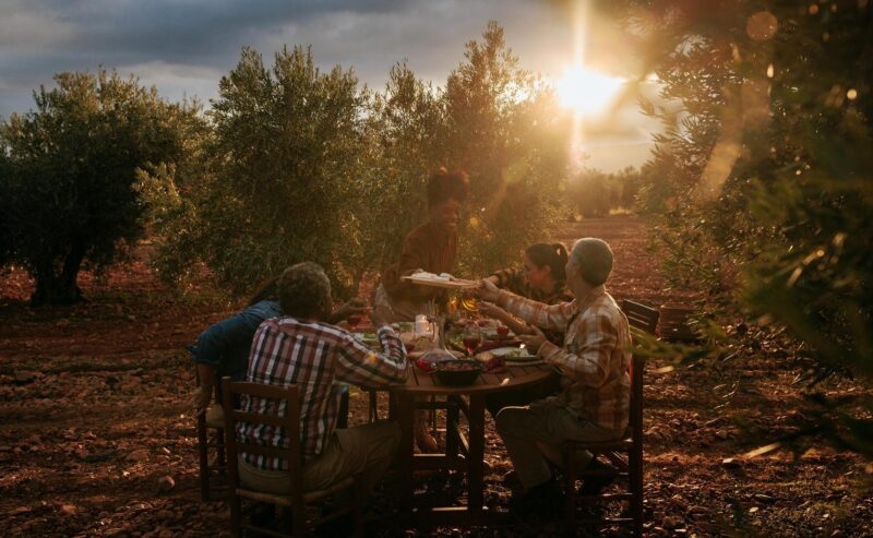 Group of people sitting at an outdoor wooden table at sunset, with one person standing to serve food.