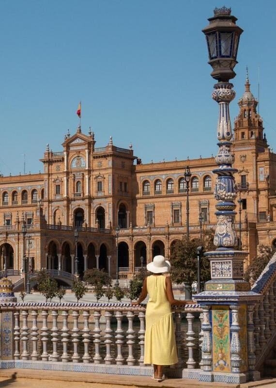 Person in a yellow dress and white hat standing on a tiled bridge looking at a large ornate historic building.