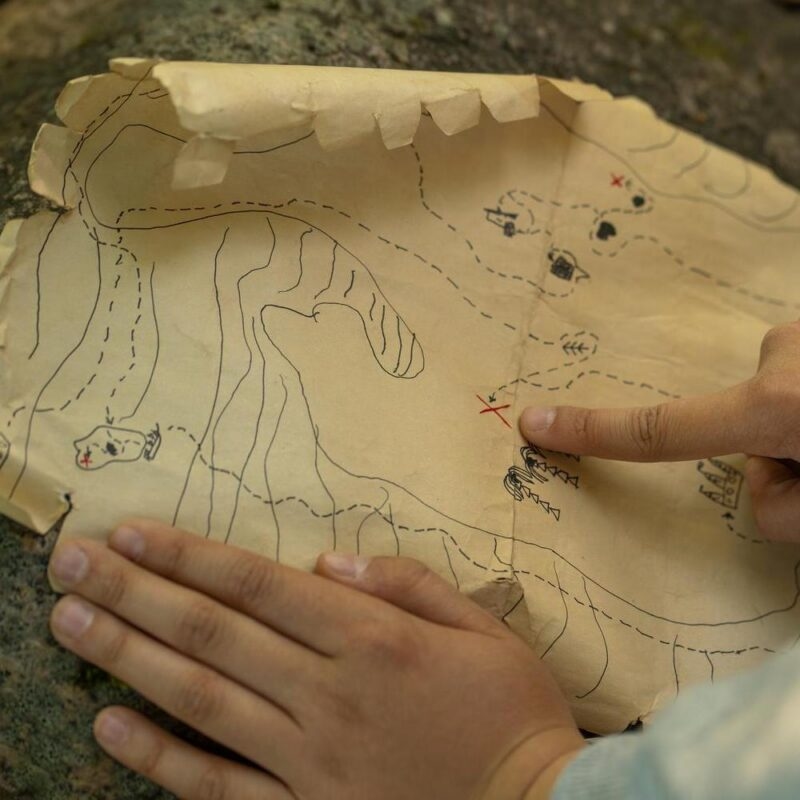 Hands holding a crinkled piece of paper showing a hand-drawn map, with one finger pointing to a red X mark.