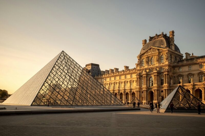 Large glass pyramid located in a paved courtyard directly in front of a grand, historic stone palace at sunset.