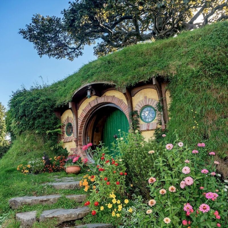 A round green door built into a grassy hill in hobiton, with stone steps and colorful flowers in the foreground.
