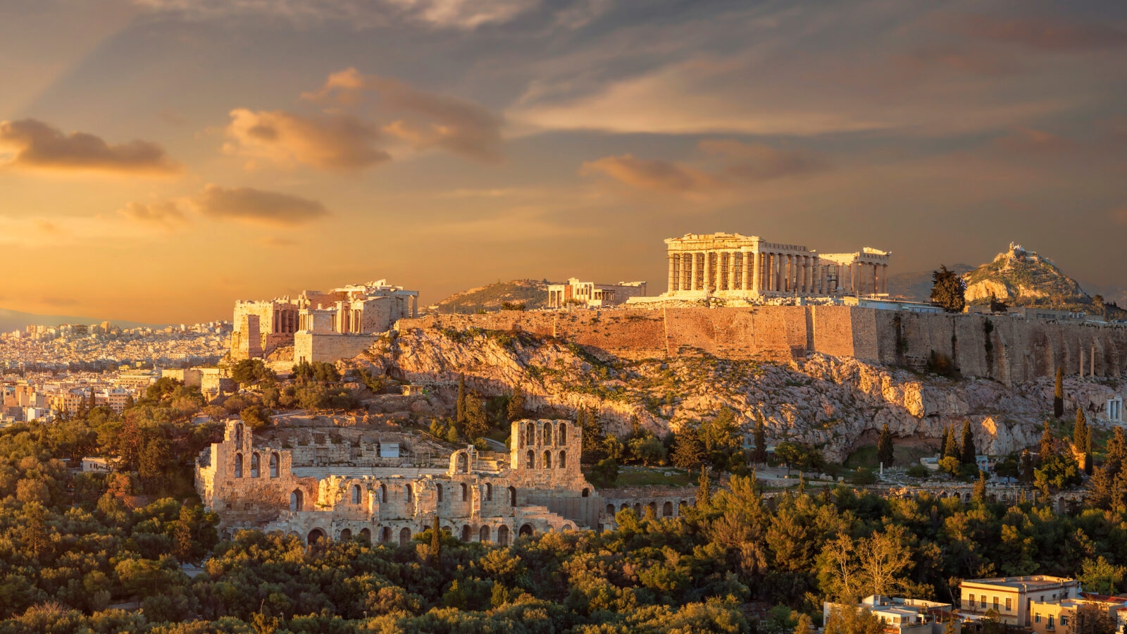 The Akropolis of athens at sunset, showing illuminated ancient temples on a rocky hill above stone ruins and green trees.