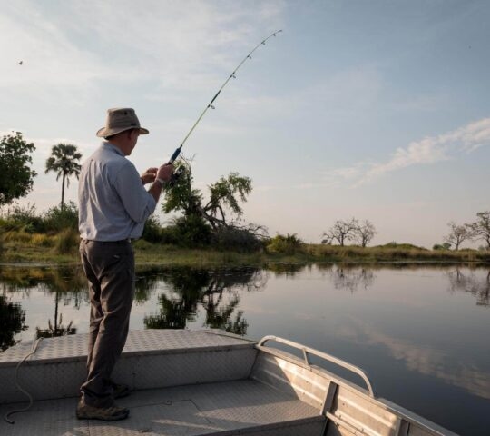 A man standing on a small boat casts a fishing line into a calm river lined with trees.