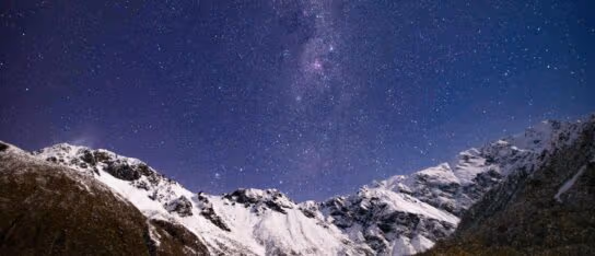 A snow-covered mountain range standing under a clear night sky full of stars.