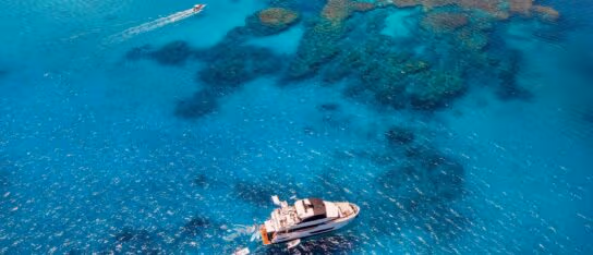 Aerial view of a white yacht anchored in clear turquoise ocean water beside visible coral reefs.