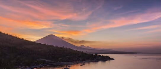 The Bali coastline under a colourful sunset sky reflected in the quiet ocean water below.