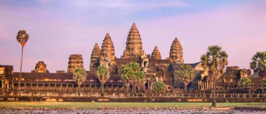 Stone towers of an ancient temple complex reflect in a pool of water with palm trees under a dusk sky.